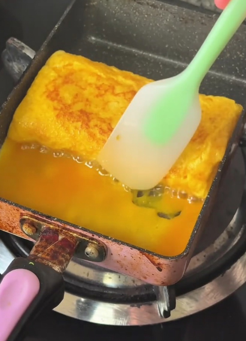 Una sartén rectangular con un rollo de huevo dorado siendo doblado con una espátula de silicona.
