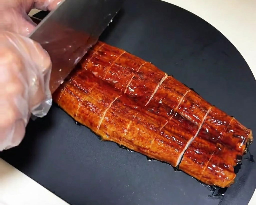 Un filete de anguila previamente asado siendo cortado en piezas rectangulares uniformes sobre una tabla de madera oscura.
