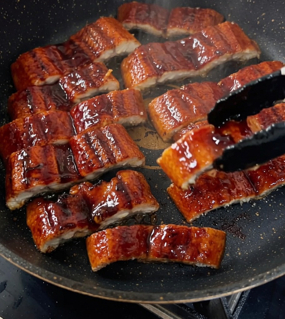 Trozos de anguila a la parrilla siendo glaseados en una sartén con una salsa oscura y rica.