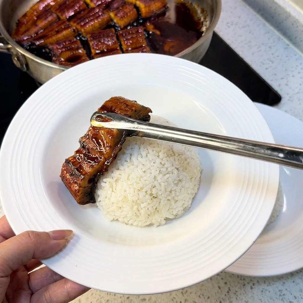 Pinzas colocando un trozo de anguila a la parrilla sobre una cama de arroz blanco en un tazón blanco, con tiras de huevo preparadas al lado.