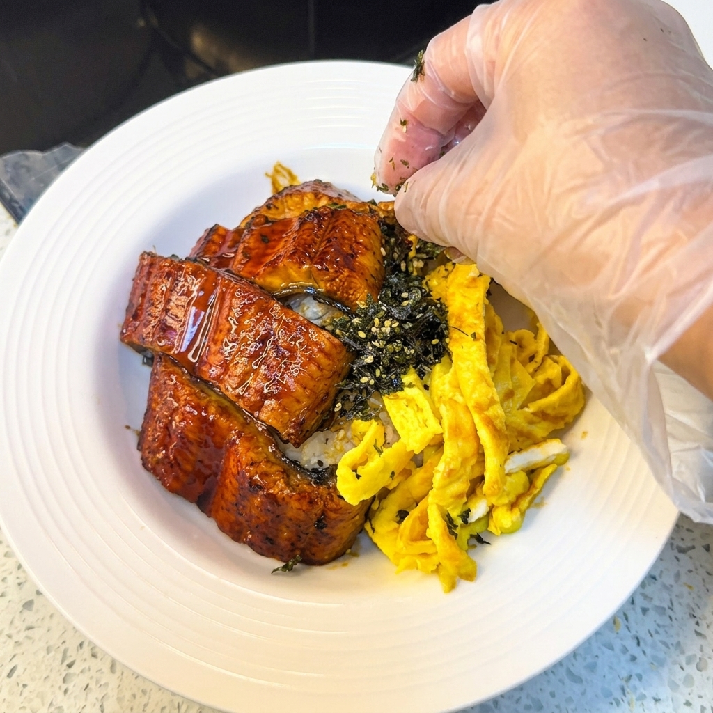Una mano con un guante de plástico espolvoreando nori rallado y semillas de sésamo sobre un tazón de anguila a la parrilla y arroz con tiras de huevo.
