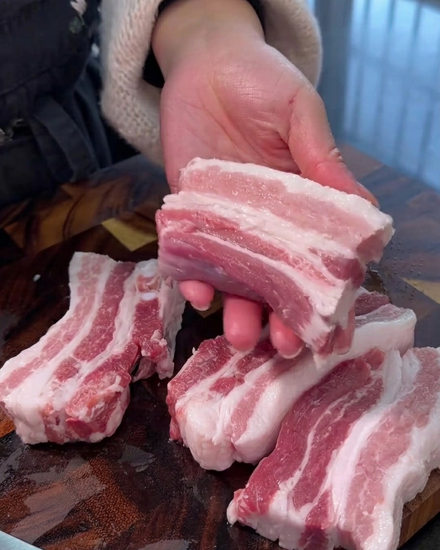 Panceta de cerdo cruda cortada en tiras gruesas y uniformes sobre una tabla de cortar de madera.