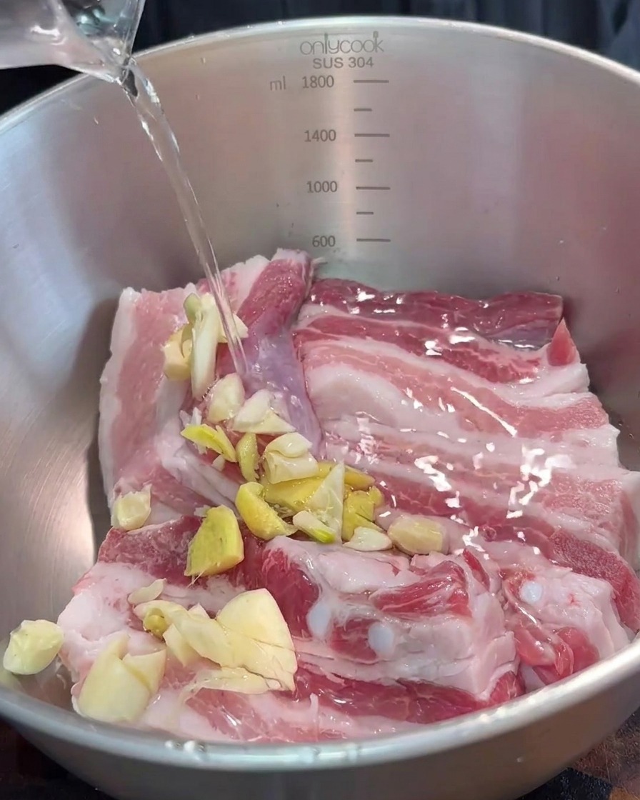 Un bol que contiene tiras de panceta de cerdo siendo sazonadas con aromáticos picados y salsa de tofu fermentado rojo.