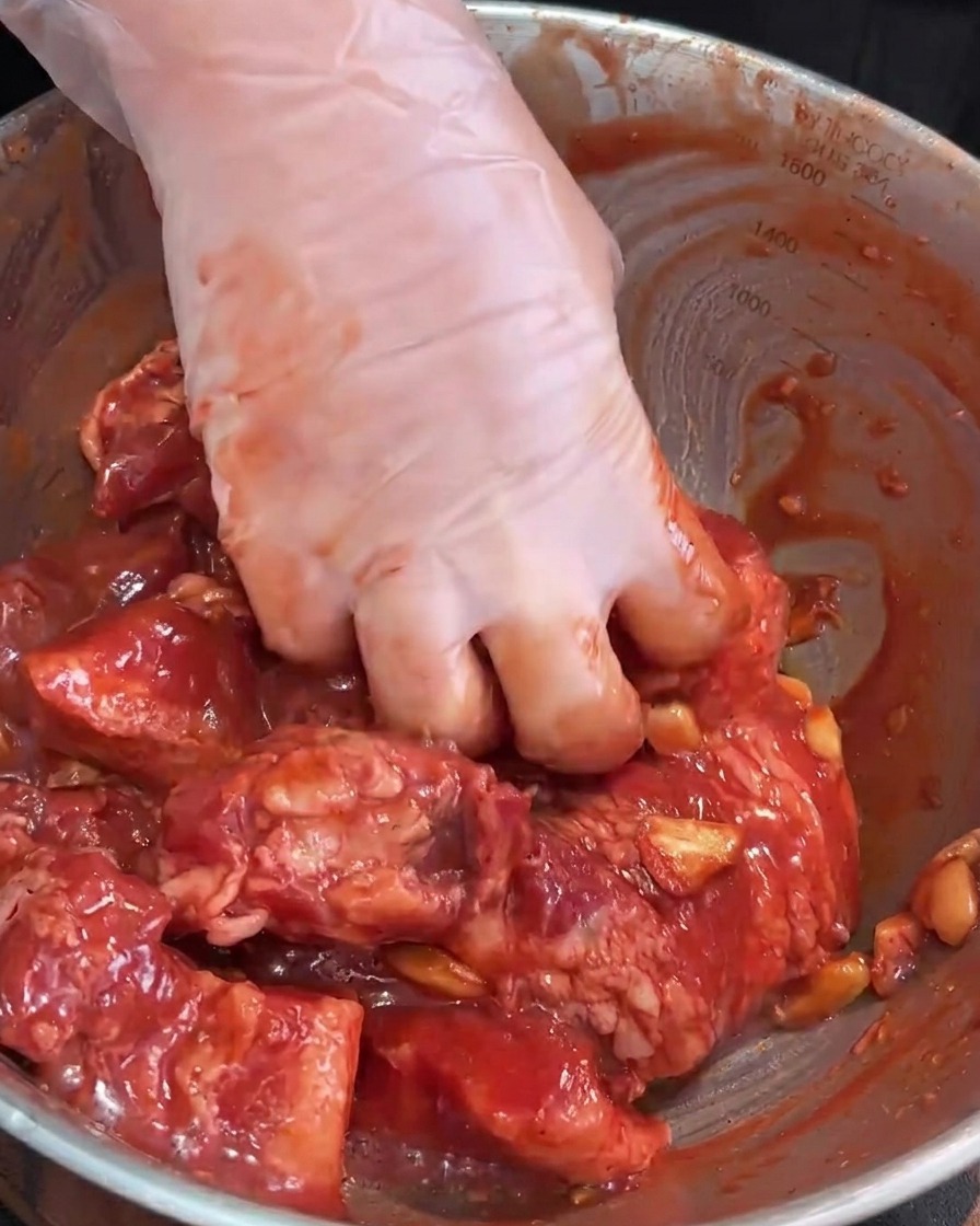 Manos con guantes masajeando el marinado en la panceta de cerdo cruda dentro de un bol de mezcla de acero inoxidable.