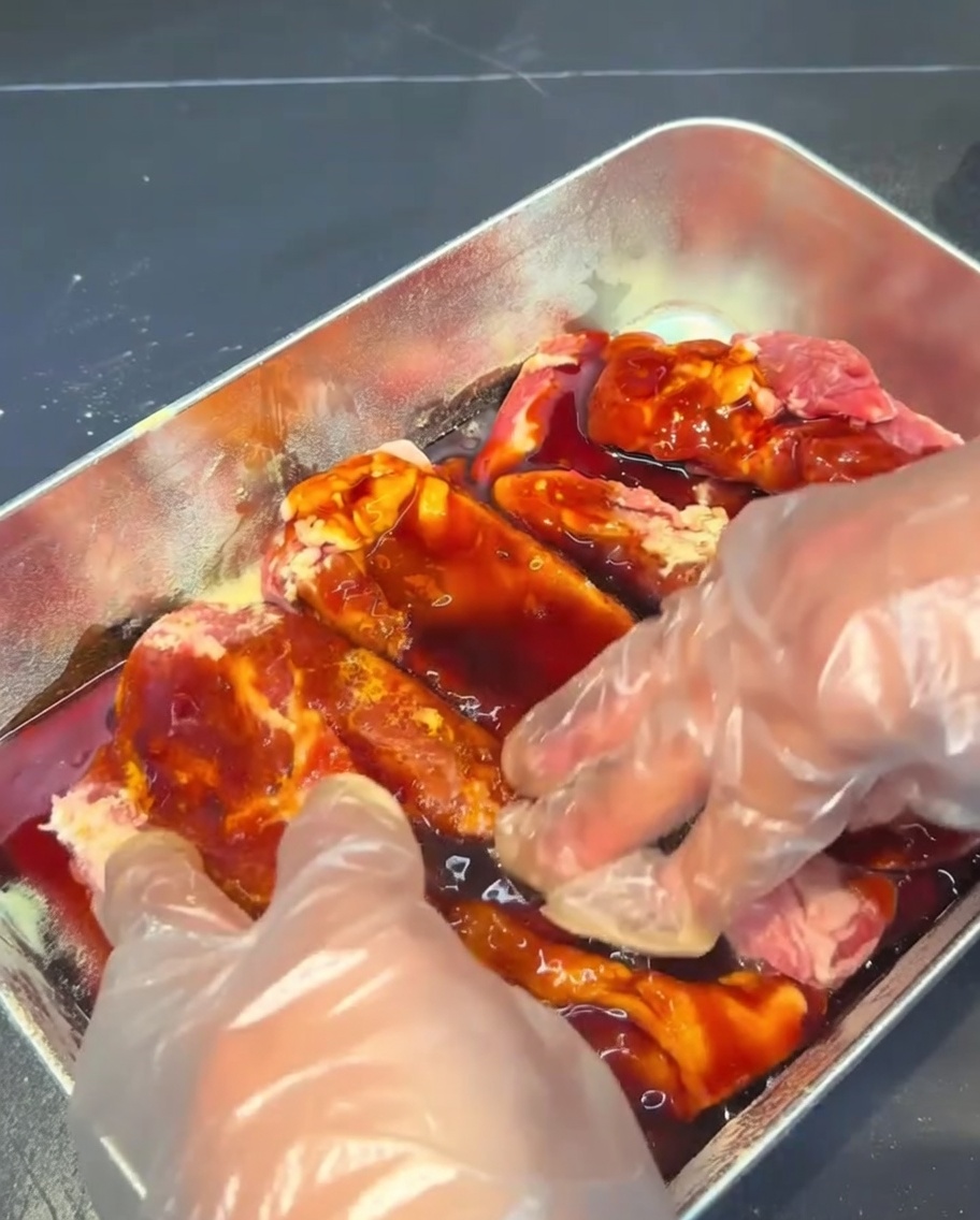 Costillas siendo masajeadas con el marinado oscuro en un recipiente metálico.
