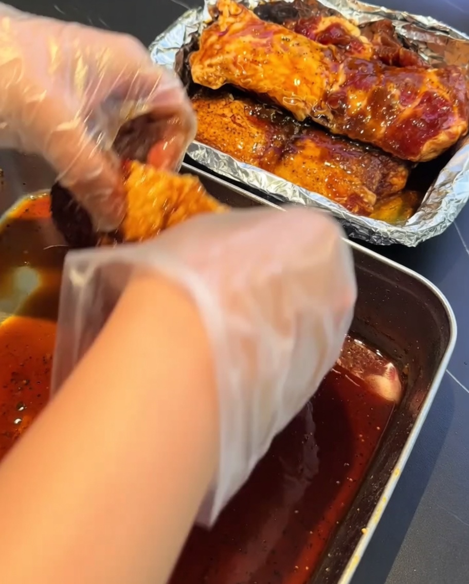 Manos con guantes transfiriendo las costillas a un recipiente forrado con papel aluminio.