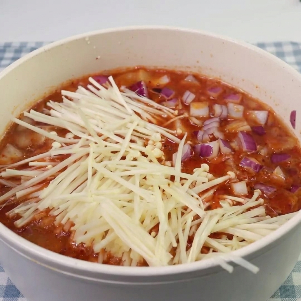 Champiñones enoki y trozos de spam siendo añadidos a la sopa de kimchi hirviendo.