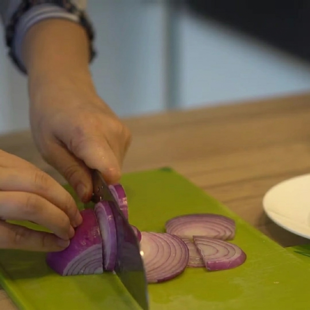 Cebolla morada siendo cortada en tiras finas sobre una tabla de cortar verde.