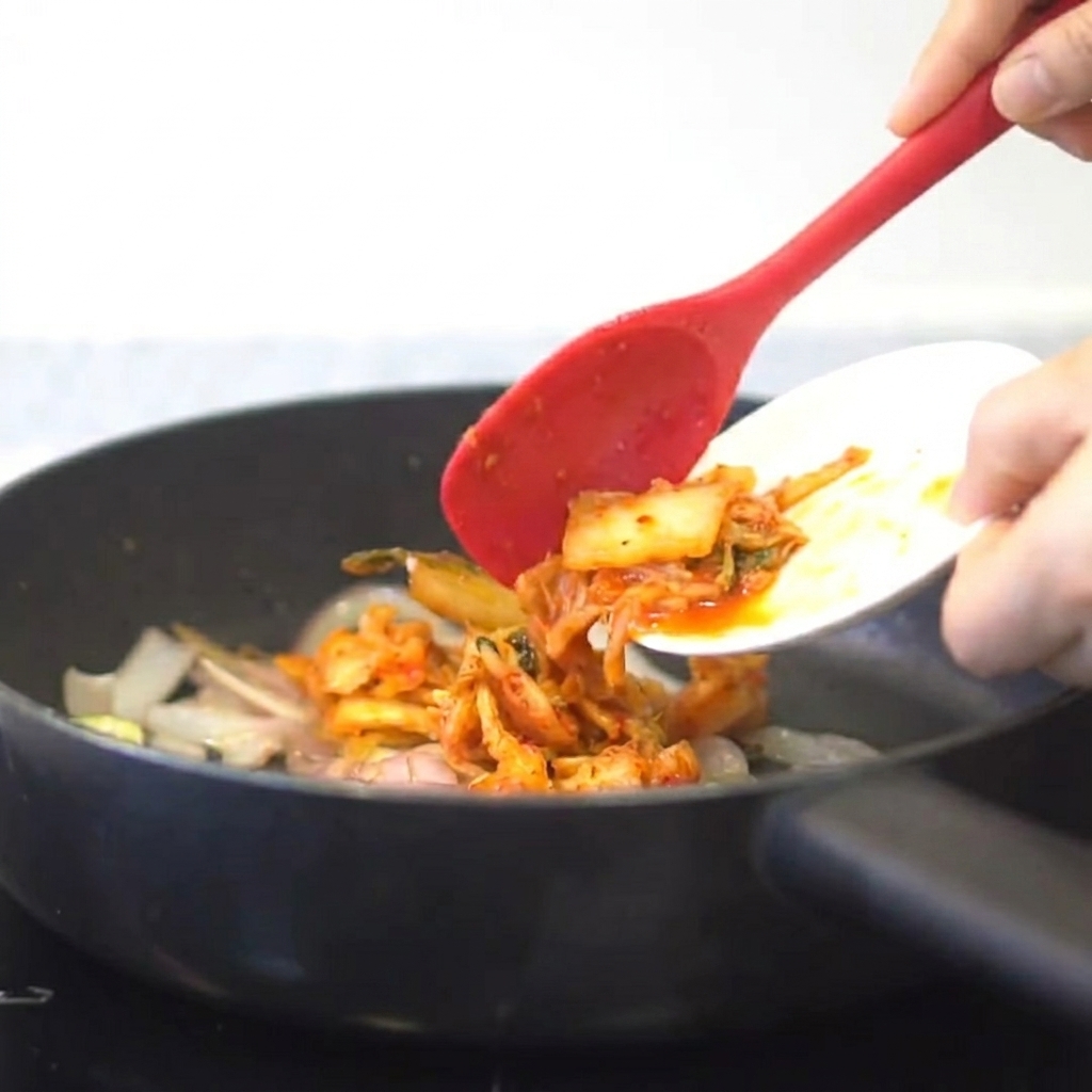 Kimchi siendo añadido a una sartén con cebollas y ajo salteados.