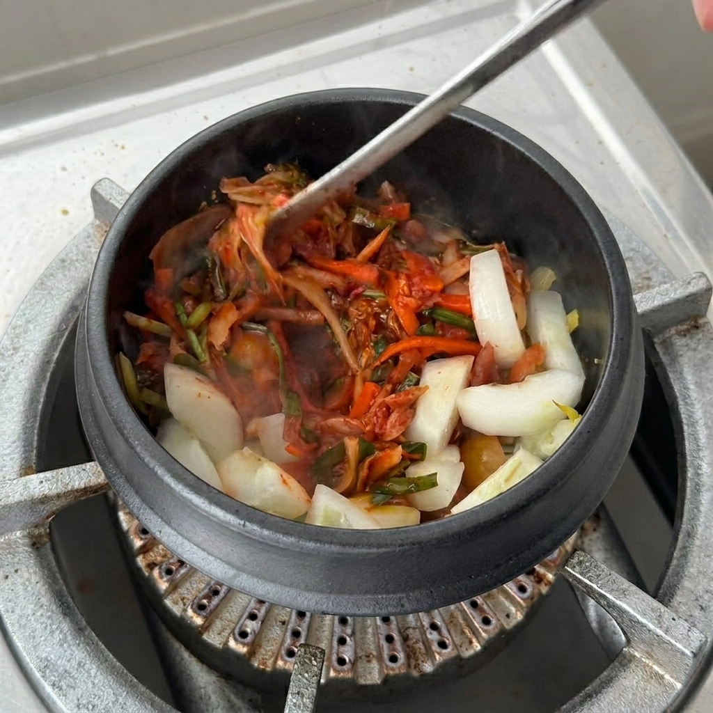 Una taza grande de base de sopa de kimchi siendo vertida en la olla de barro sobre el kimchi y las cebollas.