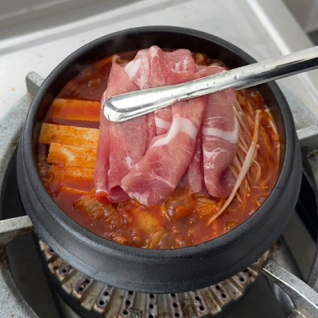 Láminas de cerdo crudo siendo colocadas sobre los ingredientes del Kimchi Jjigae que hierve a fuego lento.