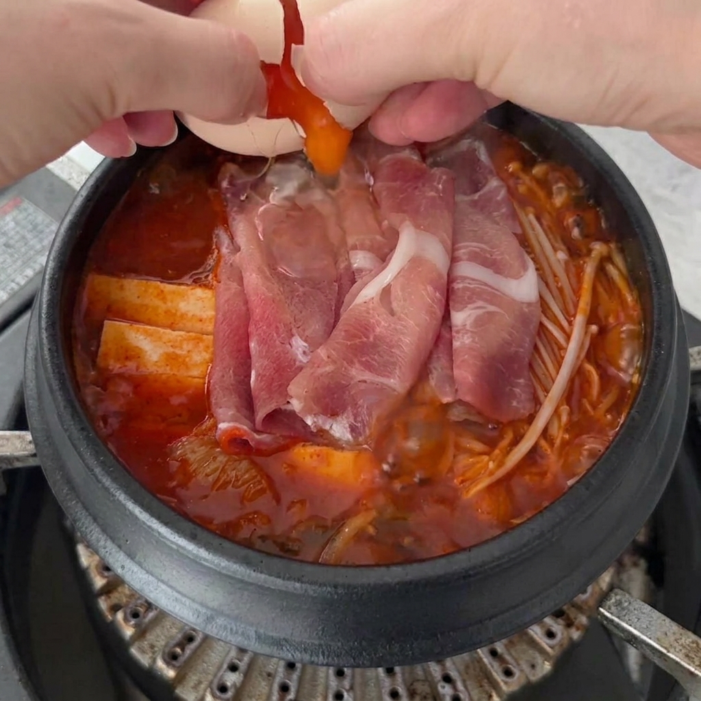 Dos huevos frescos siendo cascados sobre la superficie burbujeante del Kimchi Jjigae.