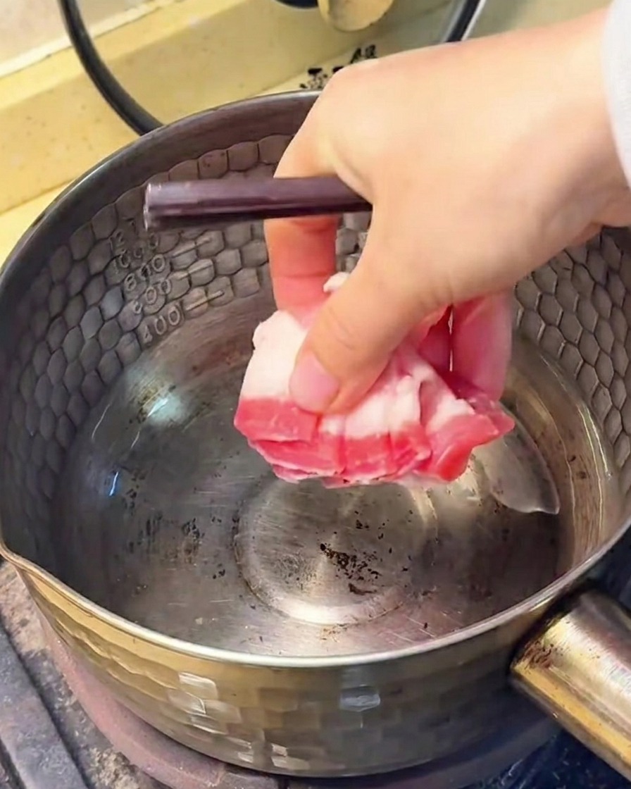 Tiras de panceta de cerdo friéndose en una olla de acero inoxidable, con la grasa visible en el fondo.