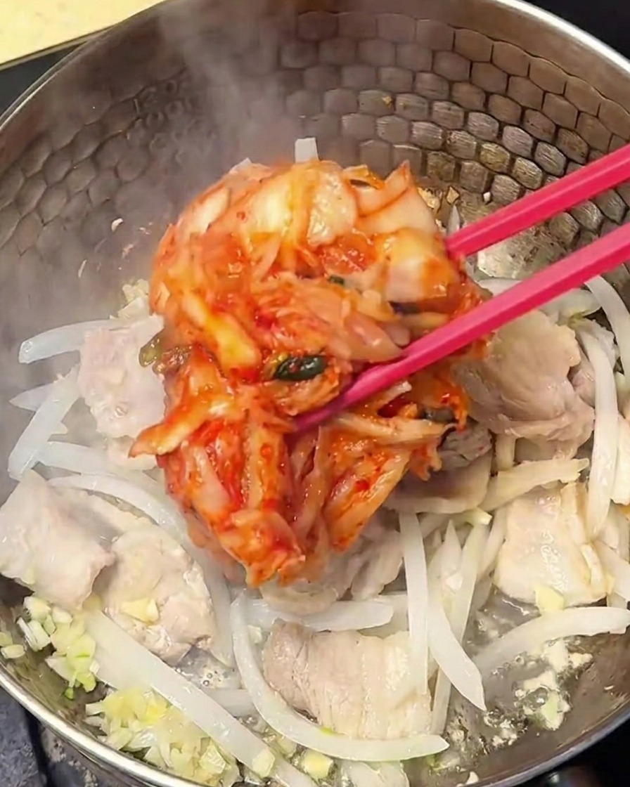 Kimchi sazonado y rojo siendo incorporado a la mezcla de cerdo y cebolla en la olla.