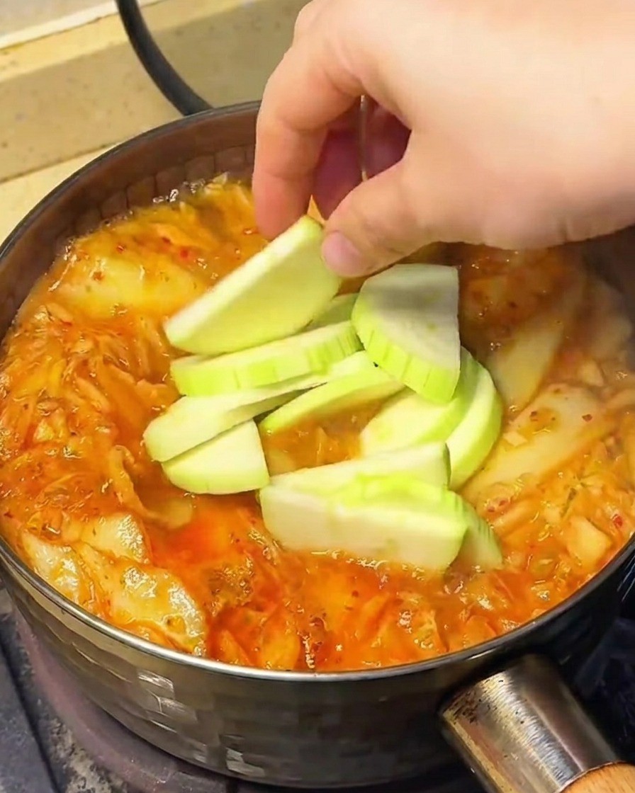 Verduras, incluyendo chayote en rodajas, brotes de soja y setas enoki, siendo añadidas a la sopa hirviendo.