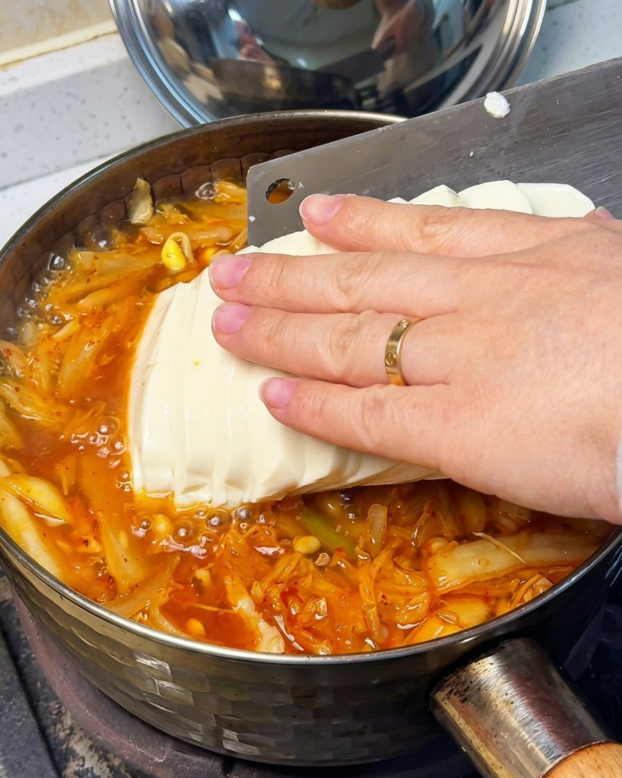 Primer plano de una mano colocando suavemente rodajas de tofu sedoso en la sopa roja hirviendo.