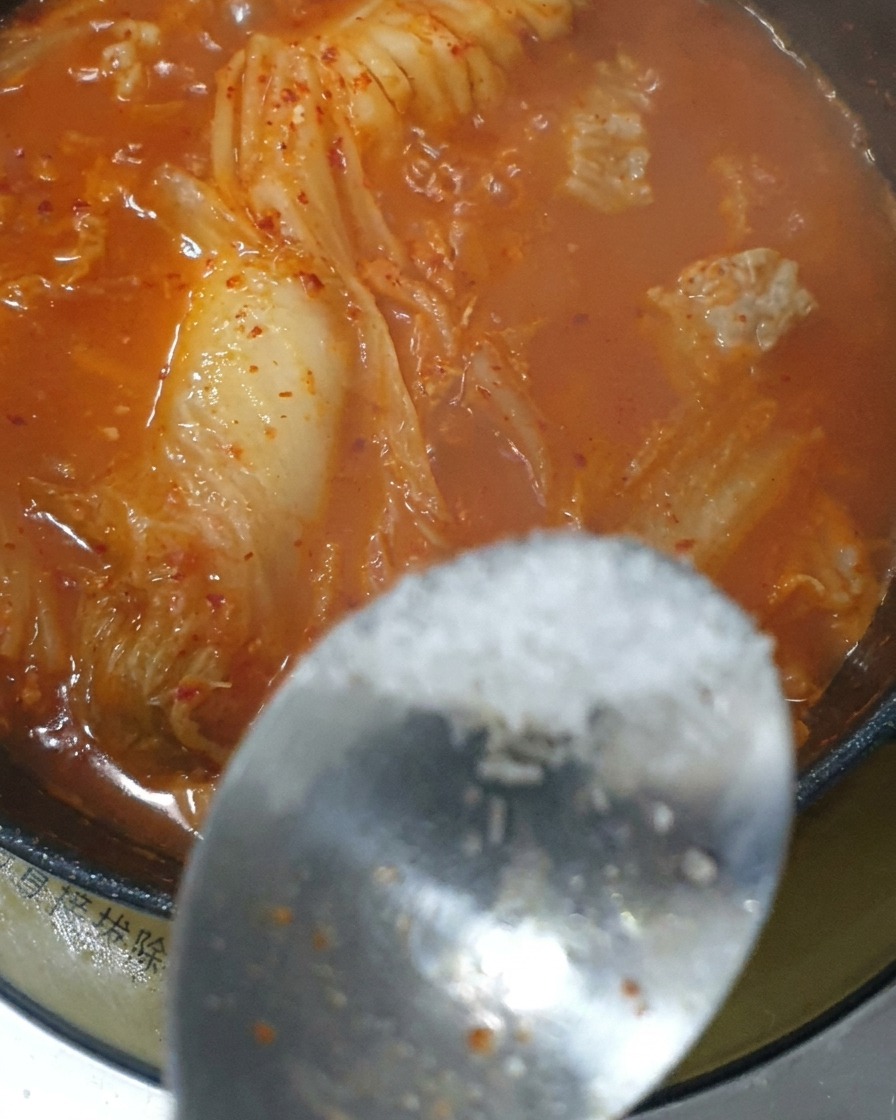 Una cucharada de condimento siendo añadida al estofado rojo de kimchi hirviendo.