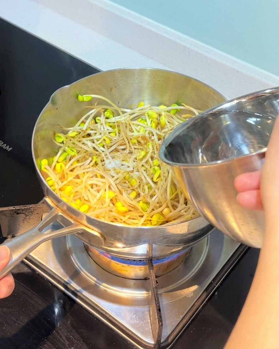 Agua siendo vertida desde un cuenco de acero inoxidable a la olla con brotes de soja y otros ingredientes.