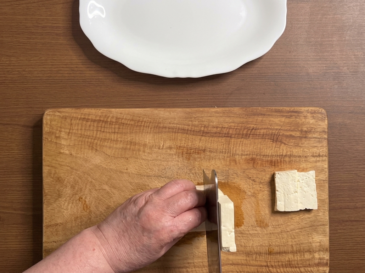 Un bloque de tofu siendo cortado en rodajas gruesas y limpias sobre una tabla de madera.