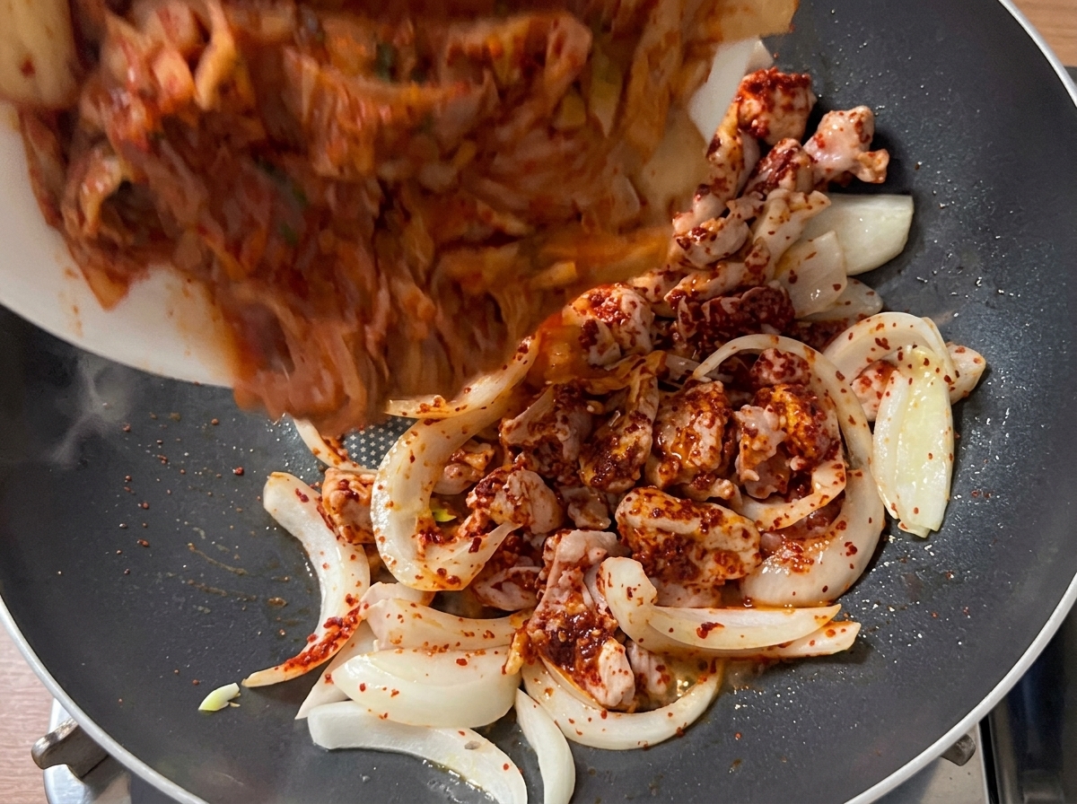 Trozos de kimchi del tamaño de un bocado siendo vertidos en la sartén con la mezcla de cerdo y cebolla dorada.
