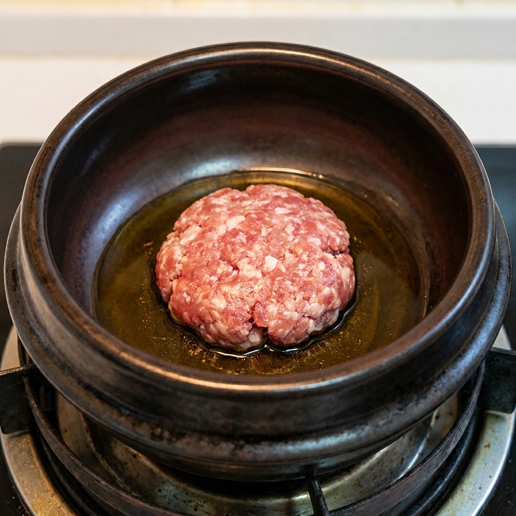 Una porción de cerdo picado dorándose en aceite de sésamo caliente dentro de una cazuela de barro oscura.