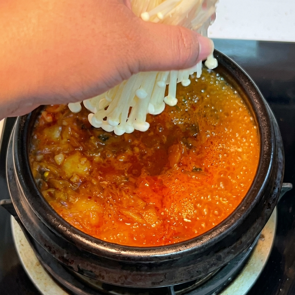 Una mano colocando un manojo de setas enoki frescas en el estofado de kimchi hirviendo.