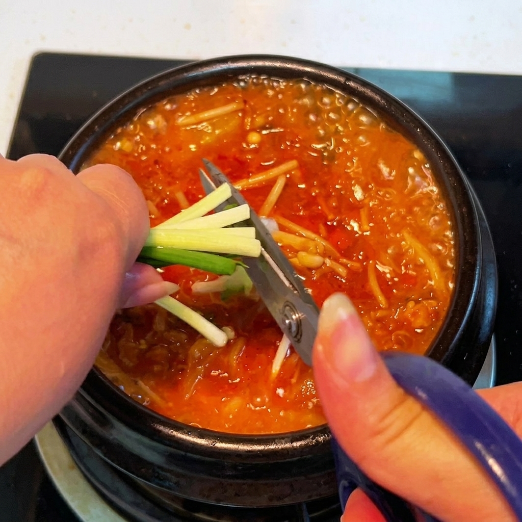 Cebolletas frescas siendo cortadas directamente sobre la cazuela de estofado de kimchi con unas tijeras de cocina.