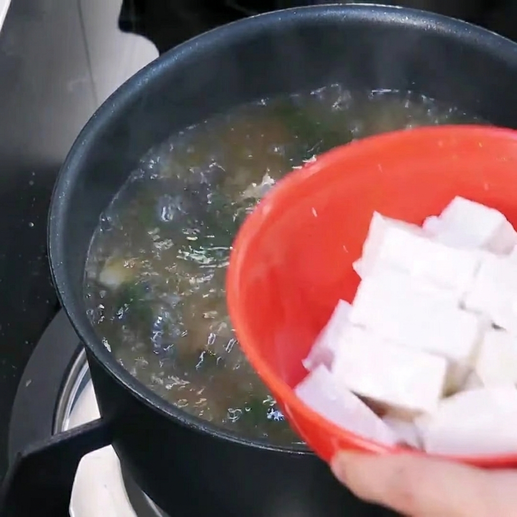 Tofu en cubos siendo añadido a una olla de sopa de pasta de soja hirviendo.