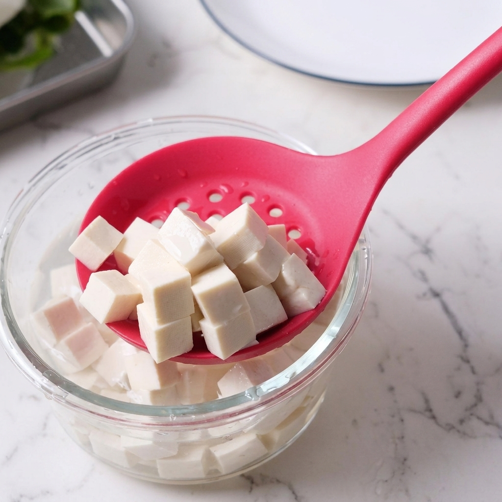Cubos de tofu siendo extraídos de un bol de agua con una cuchara ranurada rosa.