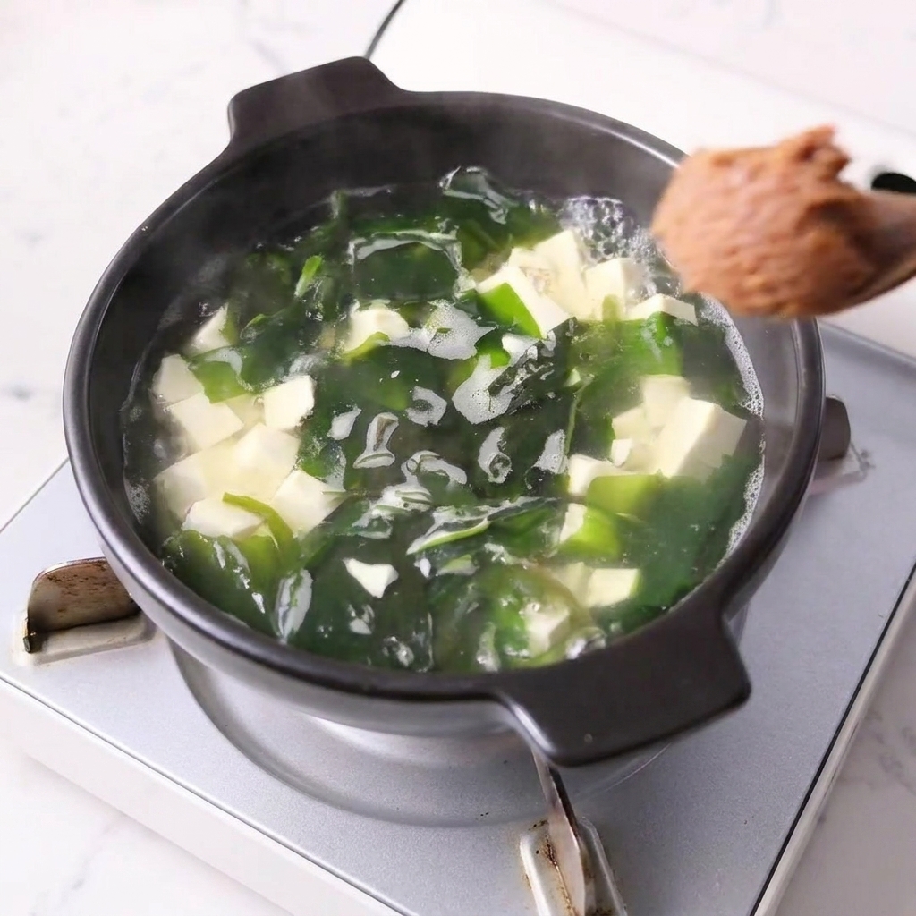 Una cucharada de pasta de miso siendo incorporada en una olla con tofu y wakame.