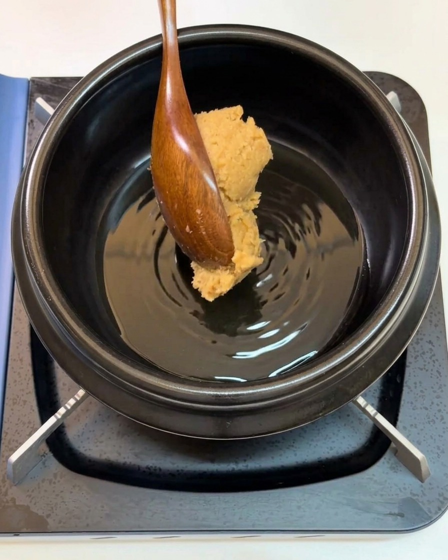 Una cuchara de madera añadiendo una cucharada de pasta de soja en una olla.
