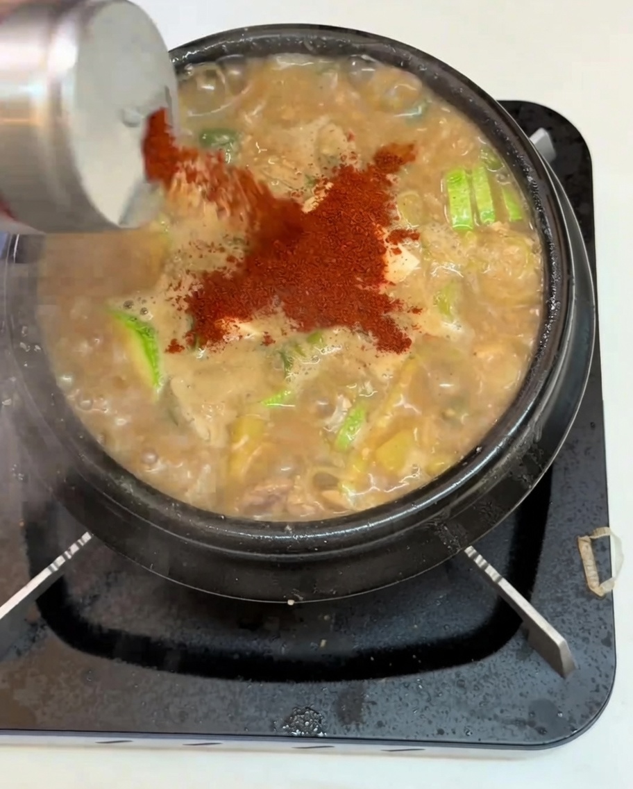 Un primer plano de chile en polvo siendo espolvoreado desde un recipiente sobre una olla de estofado de pasta de soja hirviendo, con tofu, calabacín y verduras.
