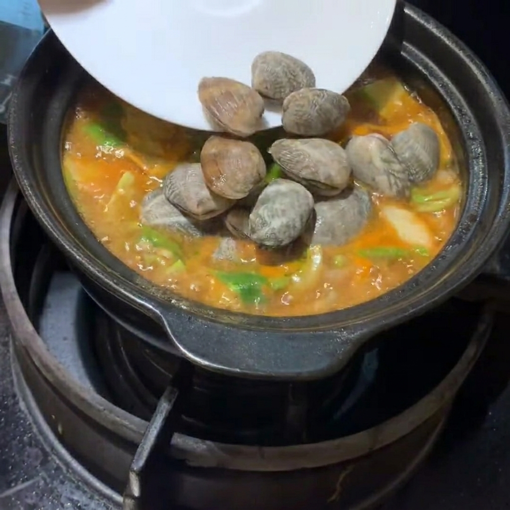 Almejas frescas y cubos de tofu suave siendo colocados cuidadosamente en el estofado.