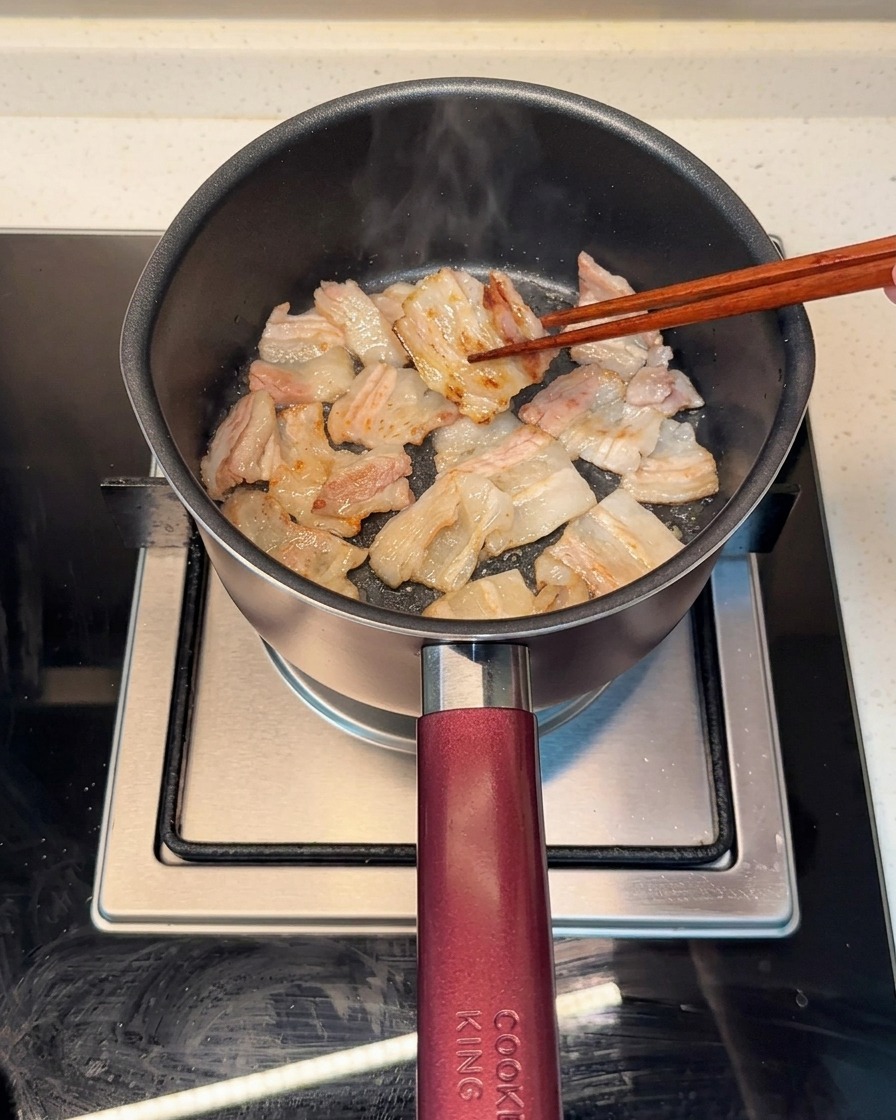 Rebanadas de panceta dorándose uniformemente en una olla caliente, con la grasa renderizada visible.