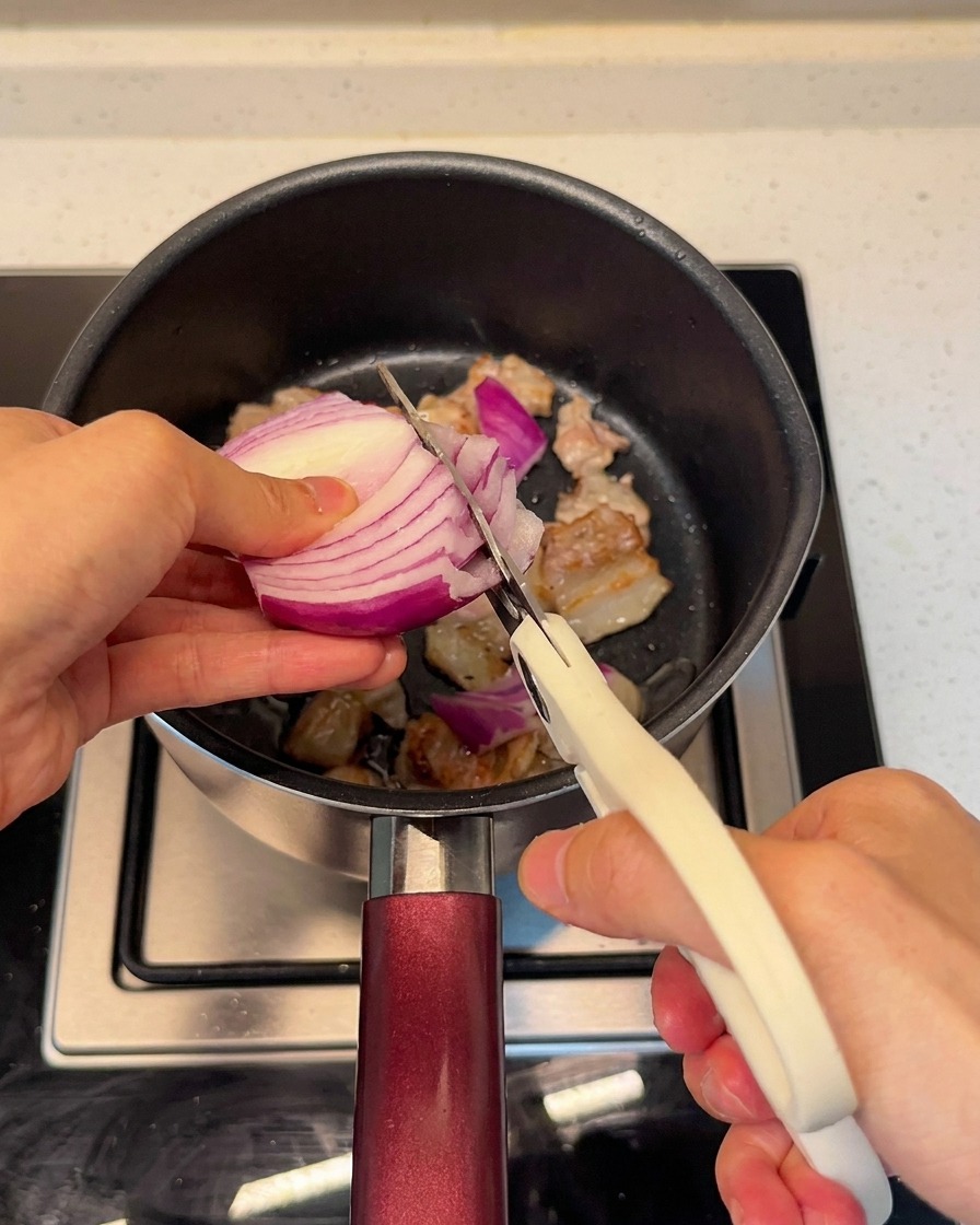 Cortando cebolla roja fresca directamente en la olla con la panceta usando tijeras de cocina.