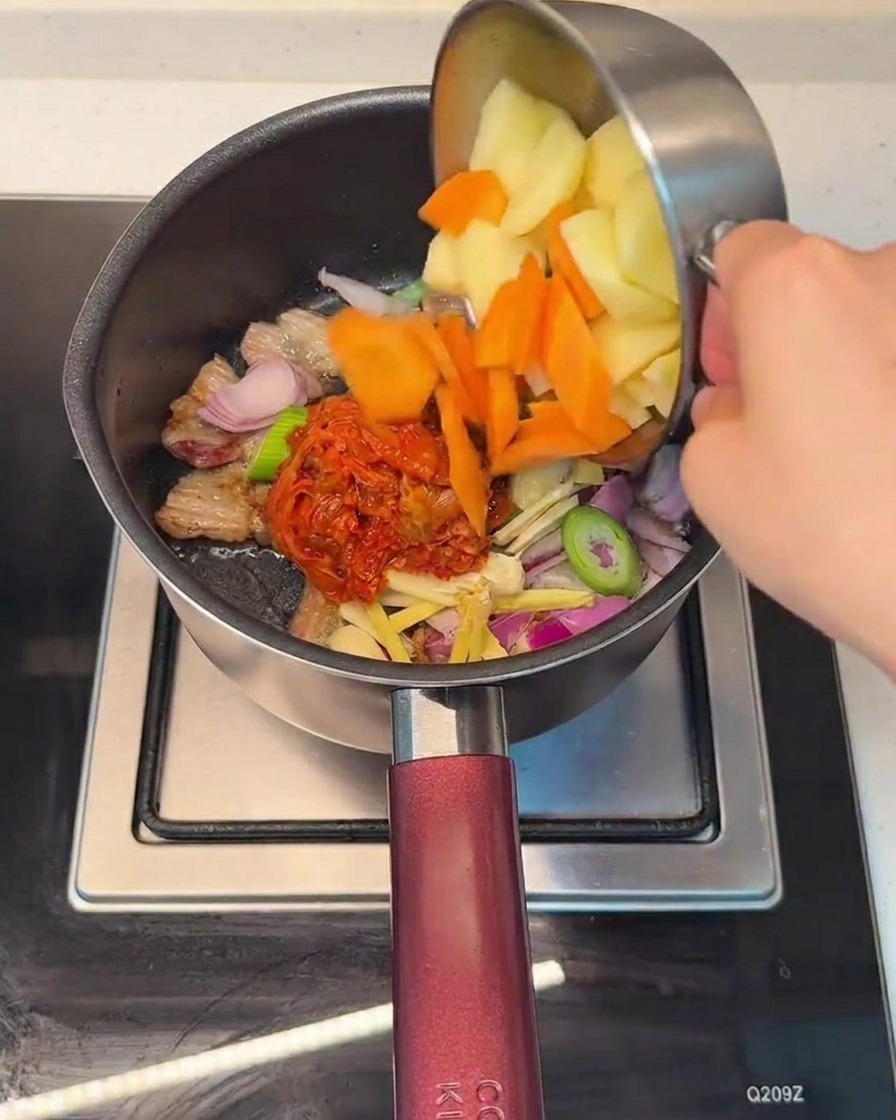 Añadiendo zanahorias y patatas picadas en la olla que contiene la mezcla de cerdo y kimchi.