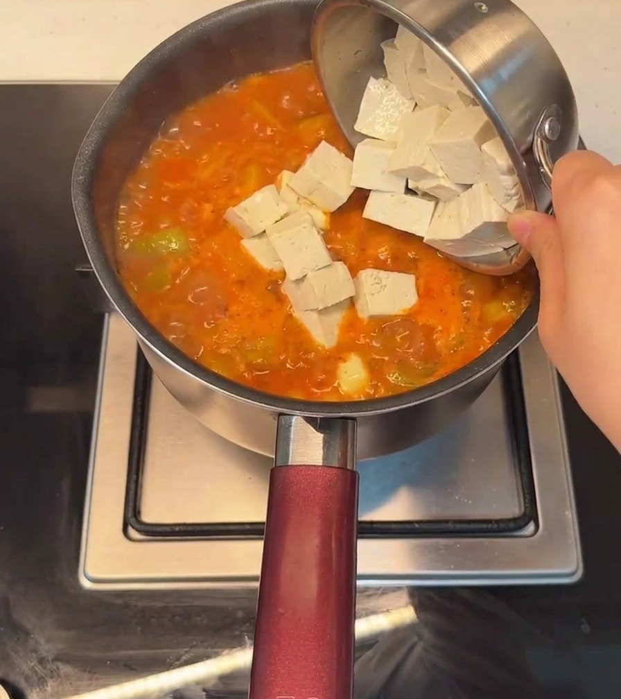 Tofu en cubos siendo añadido a una olla de estofado picante de estilo coreano burbujeante y rojo.