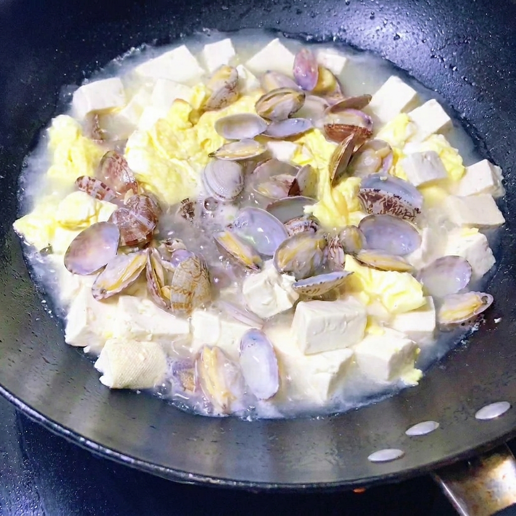 Verduras de hoja verde siendo añadidas a una sartén con estofado de almejas, tofu y huevo.