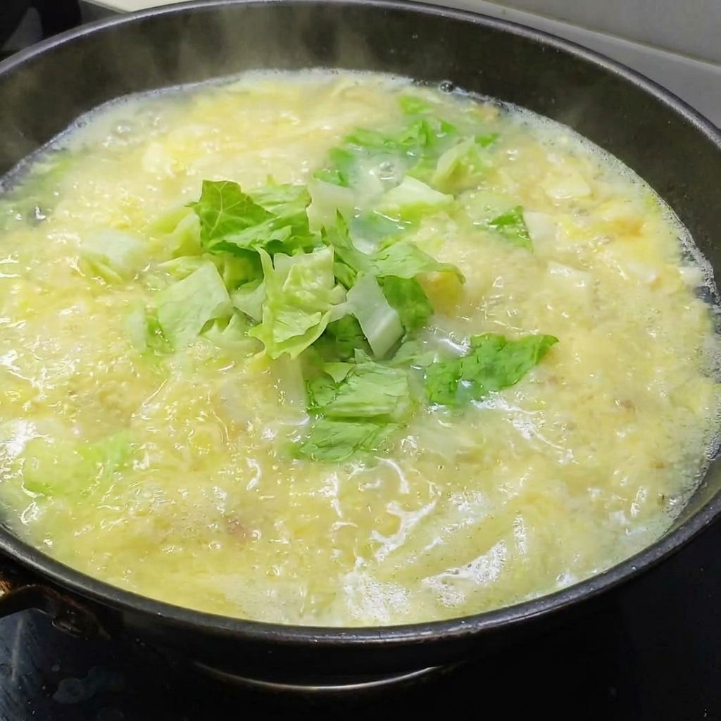 Hojas de lechuga fresca siendo añadidas a una olla de sopa dorada de huevo salado.