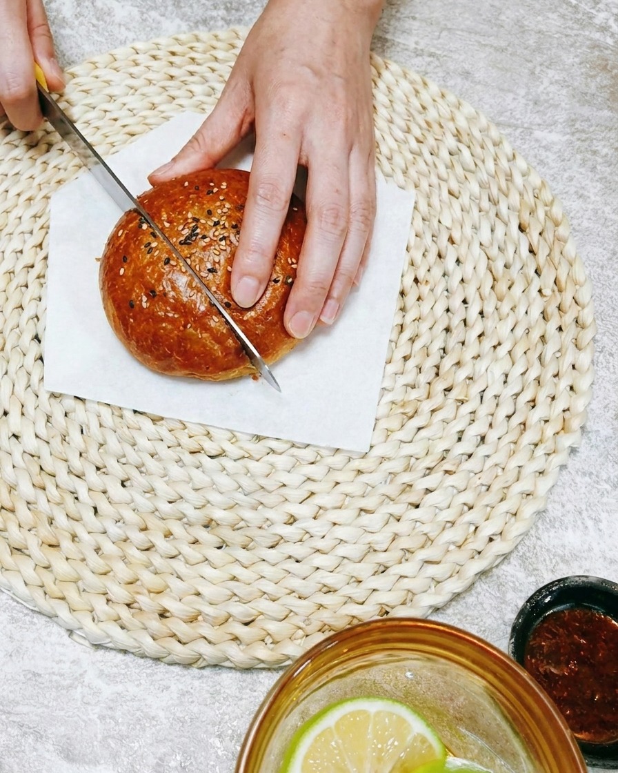 Primer plano de una mano usando un cuchillo para cortar un pan de hamburguesa suave sobre una esterilla de mimbre.