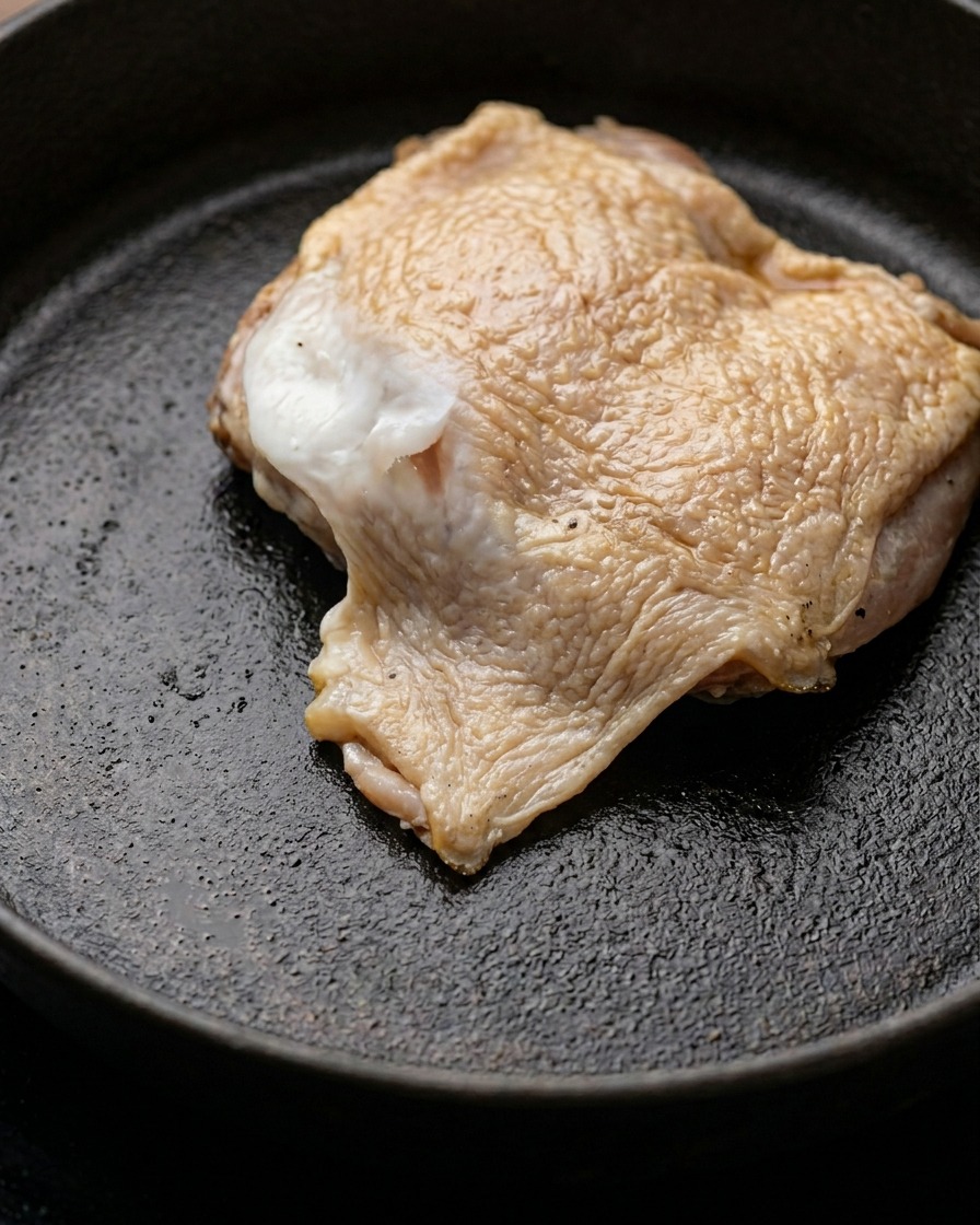 Una pieza de carne de muslo de pollo marinada dorándose en una sartén con mantequilla derretida.