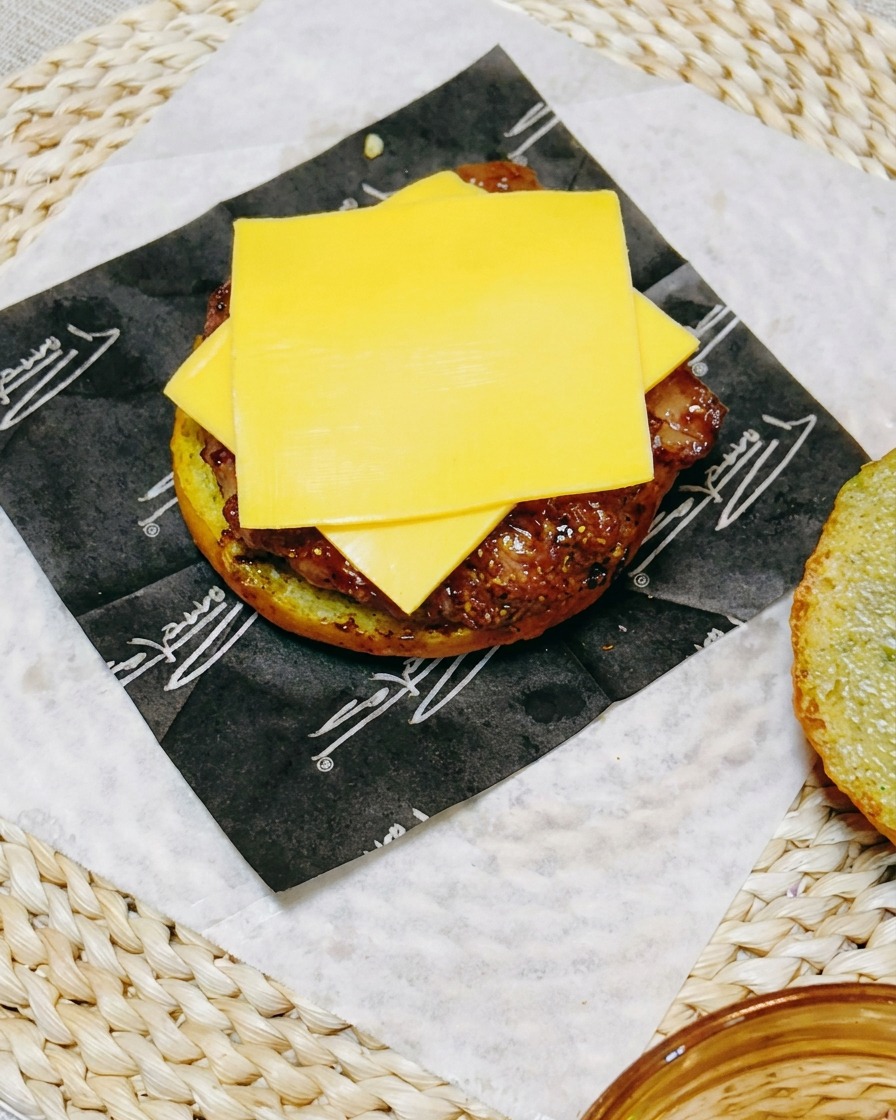 Primer plano del montaje de la hamburguesa mostrando una rebanada de queso sobre el filete de pollo, cubierto con rodajas de pepino fresco.