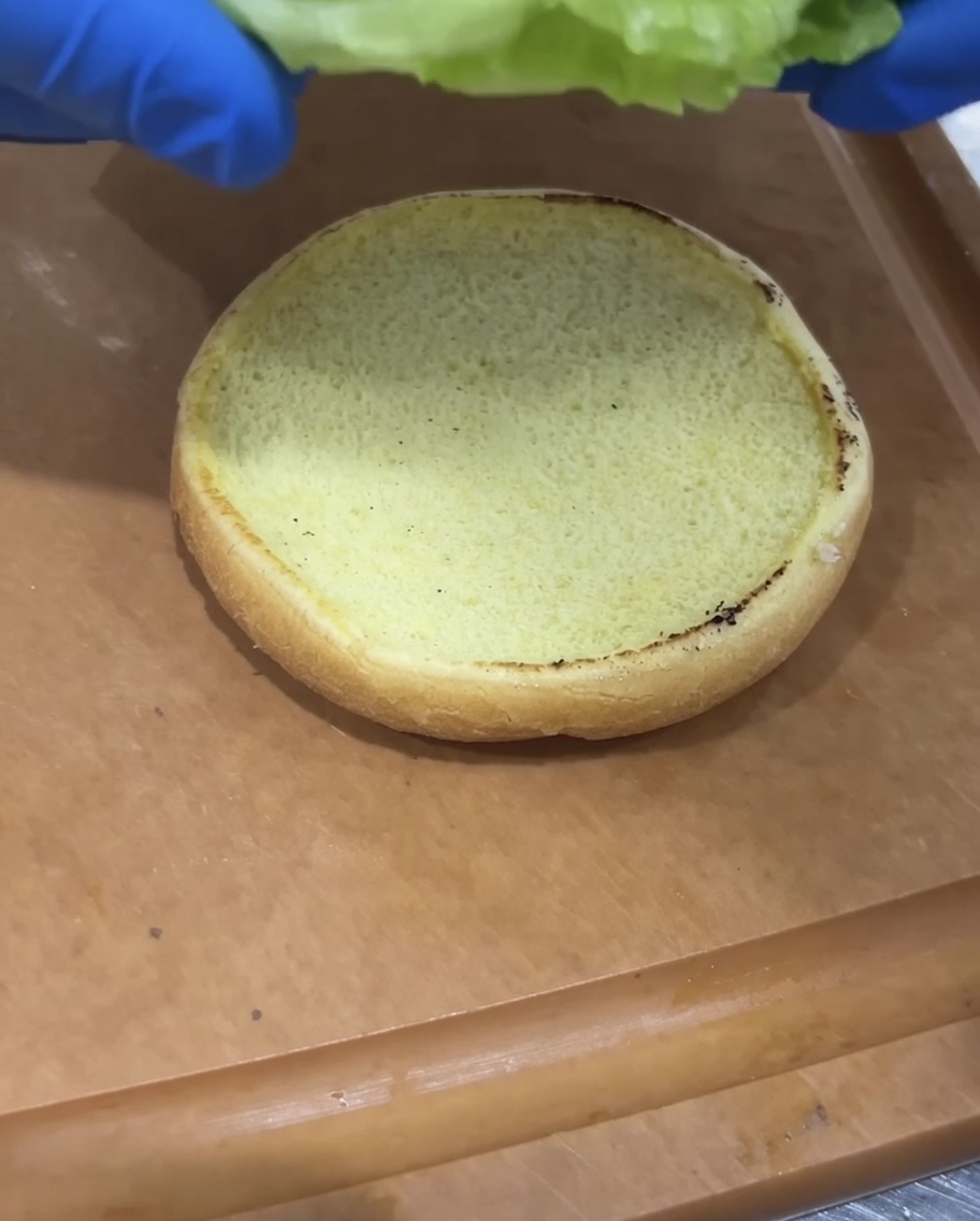 Lechuga fresca y rodajas de tomate siendo colocadas sobre un pan de hamburguesa tostado.