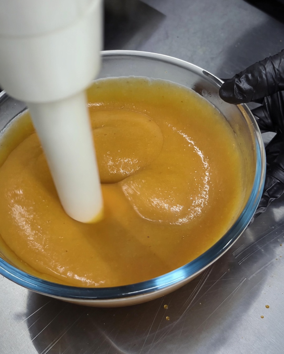 Una batidora de inmersión siendo utilizada para procesar una salsa dorada espesa en un cuenco de vidrio.