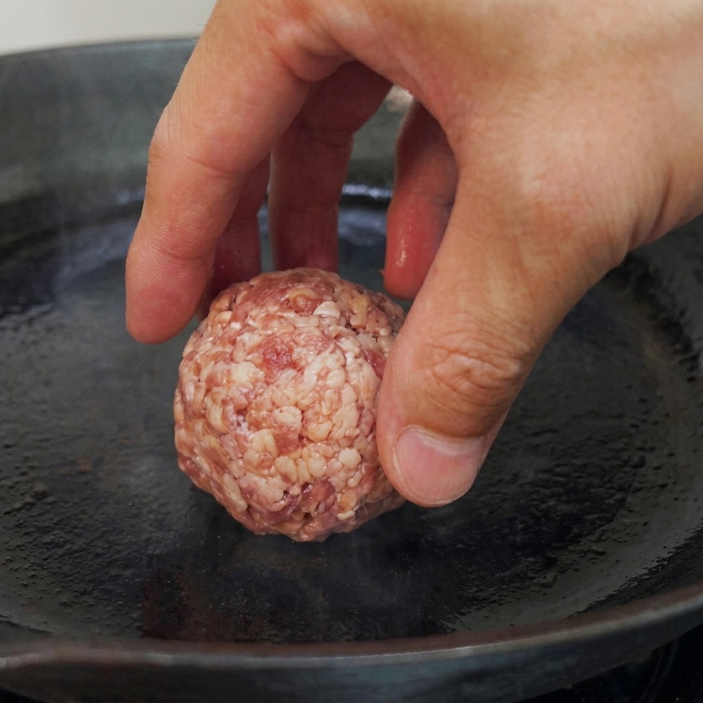 Bolas de carne cruda siendo colocadas en una sartén precalentada de fondo plano, listas para dorar.