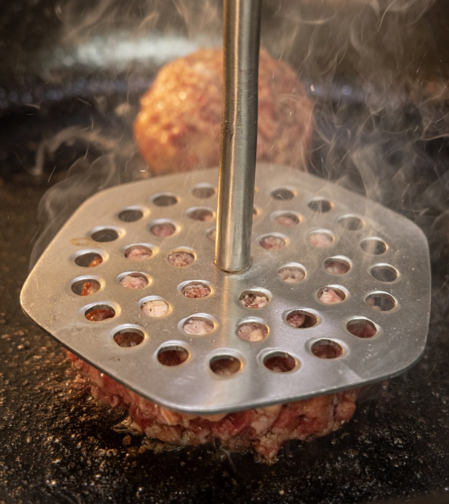 Una prensa metálica para carne siendo utilizada para aplastar bolas de carne y convertirlas en hamburguesas finas dentro de una sartén caliente.