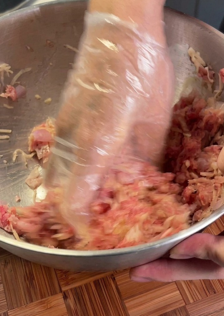 Manos con guante de plástico mezclando carne cruda y patata rallada en un cuenco metálico.