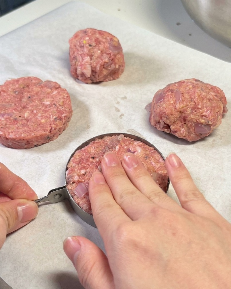 Manos usando un aro metálico para dar forma a una hamburguesa sobre papel de horno.