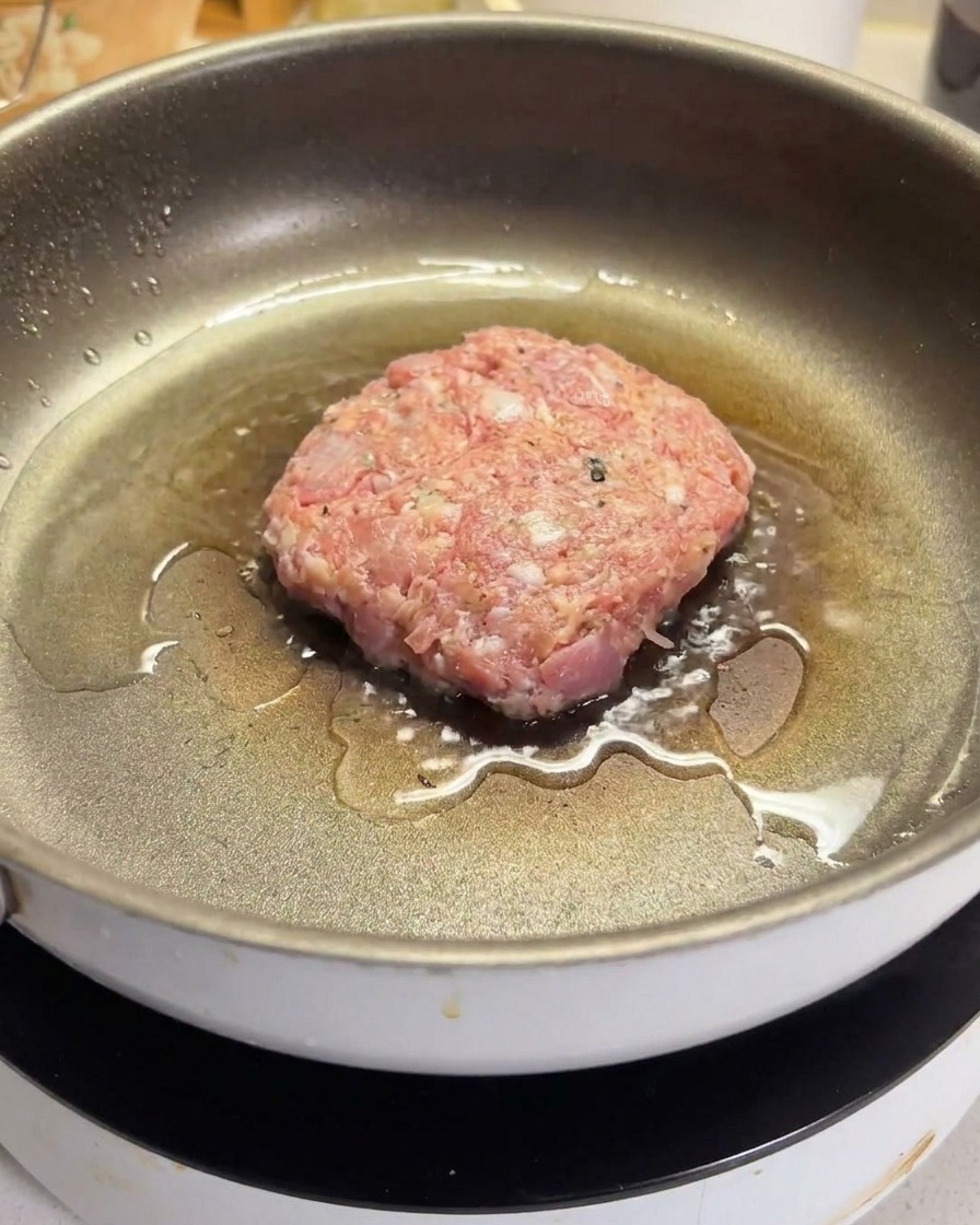Una hamburguesa de carne cruda chisporroteando en una sartén antiadherente caliente con aceite.