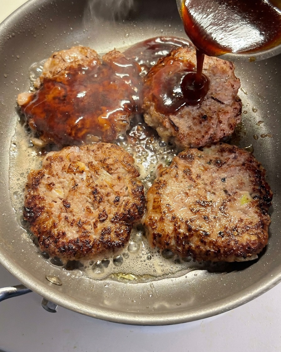 La salsa siendo vertida sobre cuatro hamburguesas dorándose en una sartén.